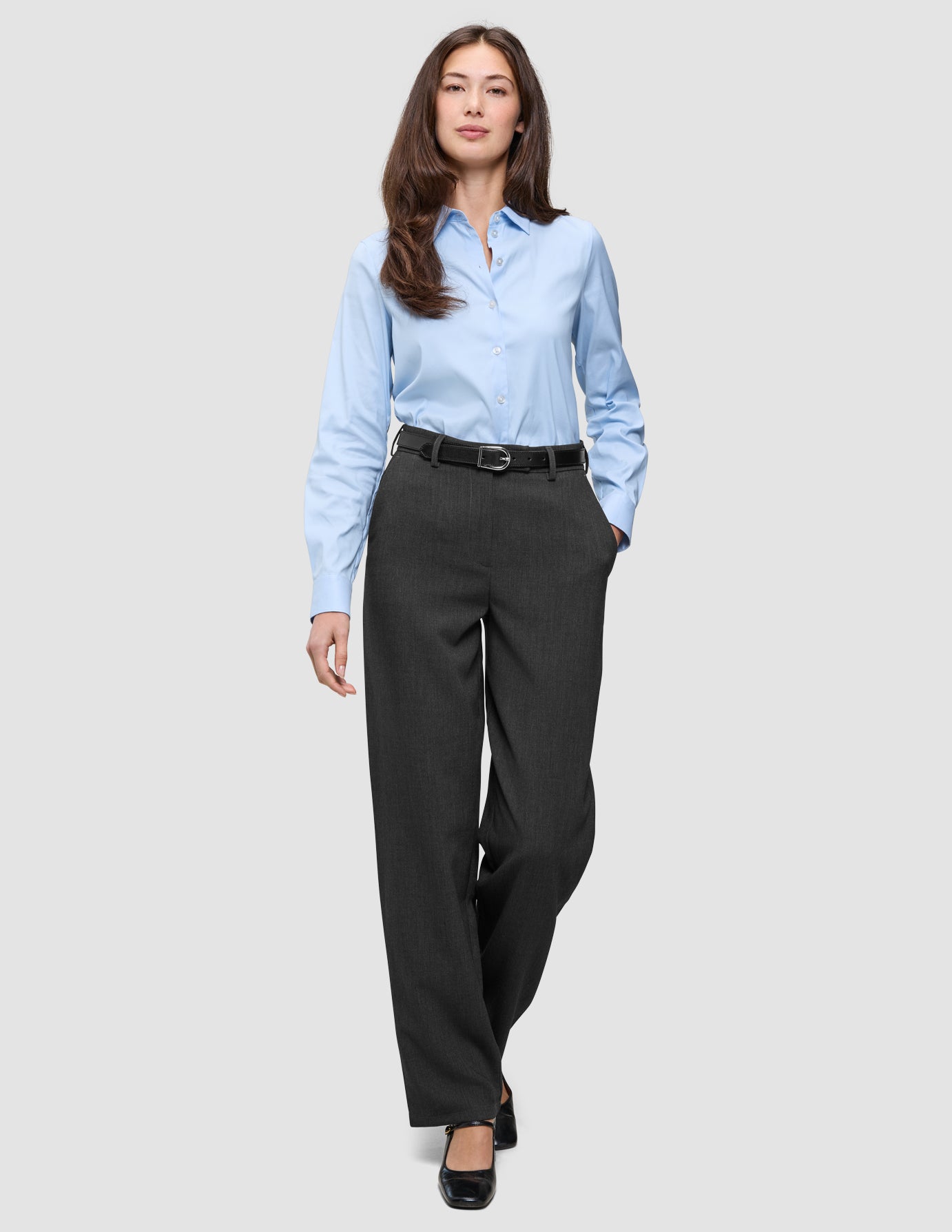 En kvinna i en ljusblå skjorta med knappar och byxor av modellen Women GO2 Pants Straight i mörkgrå melange, kombinerade med ett svart bälte.